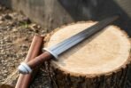 Rune engraved Viking sword laid diagonally across a tree round beside its leather covered wooden scabbard on gravel