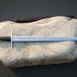 Rune engraved viking sword resting on a pale sandstone boulder beside a brown leather scabbard, with a wheel pommel, a dark leather grip and a polished blade
