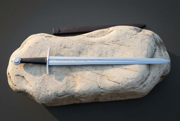 Rune engraved viking sword resting on a pale sandstone boulder beside a brown leather scabbard, with a wheel pommel, a dark leather grip and a polished blade