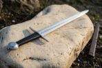 Rune engraved Viking sword with black leather grip, straight cross and wheel pommel laid on a pale boulder