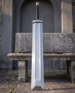 Rune etched fantasy greatsword with square guard and long black grip upright in front of a bench, scabbard laid behind