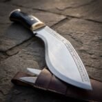 Rune etched high carbon kukri knife, close up of the brass bolster and curved blade resting on a leather sheath with small knives