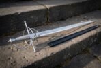 Rune etched rapier with a swept steel hilt and wire wrapped grip lying across weathered stone steps beside its black scabbard in low evening sunlight