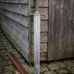 Runic guard damascus sword with olive green leather grip, brass ferrules and faceted steel pommel standing against a weathered timber wall beside its brown leather scabbard