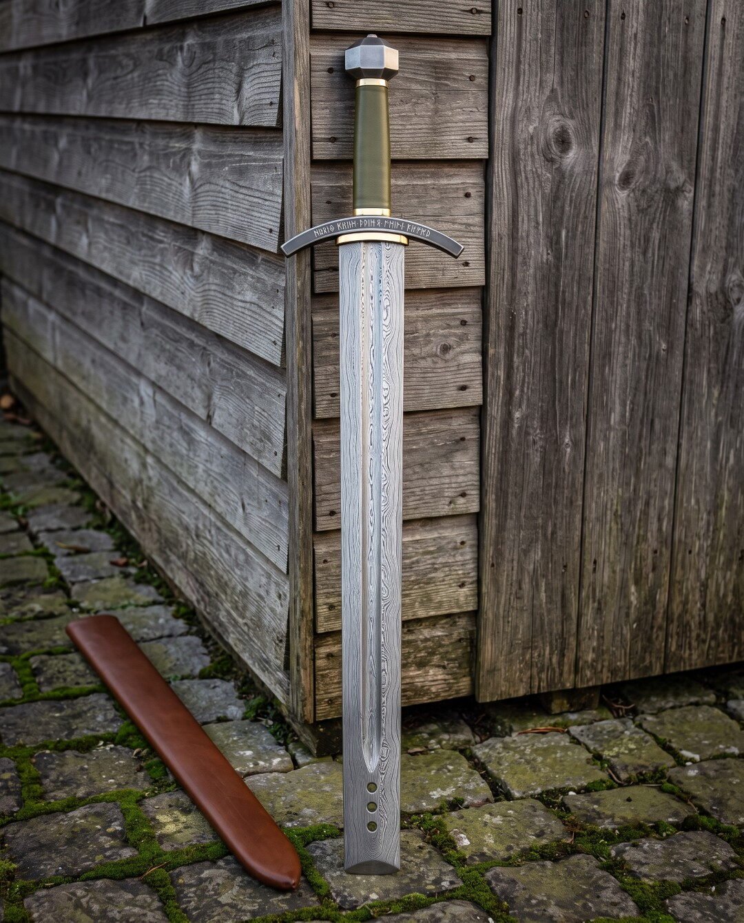 Runic guard damascus sword with olive green leather grip, brass ferrules and faceted steel pommel standing against a weathered timber wall beside its brown leather scabbard