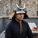 Samurai kabuto helmet with a ribbed black bowl, brass crescent crest and orange laced neck guard worn by a man outdoors