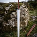 Satin polished Vegvisir arming sword with a runic compass engraved disc pommel and brown leather grip standing on moorland grass beside a lichened boulder