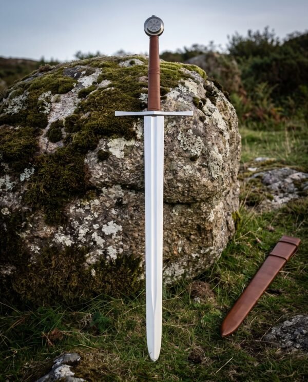 Satin polished Vegvisir arming sword with a runic compass engraved disc pommel and brown leather grip standing on moorland grass beside a lichened boulder