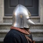 Satin steel pig faced bascinet with pointed hounskull visor, knotwork brow band and brass hinge worn in profile by a man in a dark gambeson