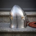 Satin steel pig faced bascinet with pointed skull, brass hinges and riveted lower edge, shown from the rear quarter on a stone step