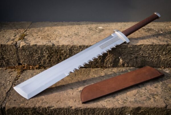 Sawback machete with an angled chisel tip and etched knotwork panel resting on a weathered stone step beside a stitched brown leather sheath in low sunlight