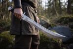 Sawback short sword with polished blade held down at the side by a man in a woodland, black grip and steel guard