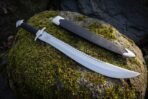 Sawback short sword with polished clip point blade, black leather grip and steel guard lying on a mossy rock by its scabbard