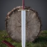 Scale etched raven pommel sword with a dragon scale blade pattern and oxblood leather grip standing against a cut log round on green grass
