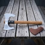 Serpent etched bearded axe in polished high carbon steel with a long sweeping edge, oak haft, blackened collar and brown leather head cover on a weathered wooden table