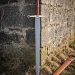 Serpent guard damascus sword with a pattern welded blade, brass quatrefoil crossguard, brown leather grip and cabochon set brass pommel leaning on an old stone wall