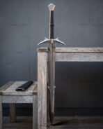Serrated fantasy sword standing point down against a weathered wooden table, twin barbed guard wings and hooked blade spurs clear against a grey concrete wall