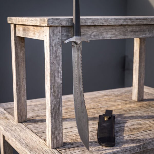 Serrated spine damascus short sword hanging point down from a weathered wooden table, showing the notched steel guard, black grip and black leather belt sheath