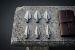 Set of six hand forged Damascus viking arrowheads with socketed bases and swept barbs laid on a weathered stone slab beside a rolled brown leather case