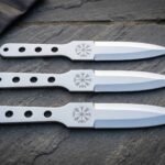 Set of three hand forged 1095 steel Viking throwing knives with skeletonised handles and Vegvisir etched blades laid on dark slate beside a leather roll