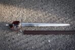 Shell guard broadsword in satin 1095 steel with a pierced hilt, deep red backing panels and a cut Helm of Awe stave lying on grey gravel beside its dark leather scabbard