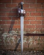 Shell guard damascus short sword with pierced steel hilt, deep red backing panels and a knotwork boss standing point down against a weathered red brick wall