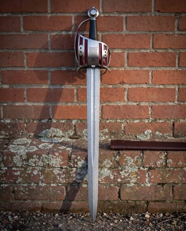 Shell guard damascus short sword with pierced steel hilt, deep red backing panels and a knotwork boss standing point down against a weathered red brick wall