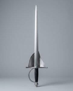 Shell guard rapier trainer with paired triangular shell plates, ball pommel, black leather grip and a ridged blade standing point up on a plain grey studio backdrop