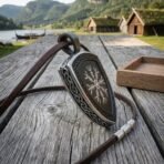 Shield shaped steel helm of awe pendant with knotwork border and brown leather cord standing on weathered grey planks by the water