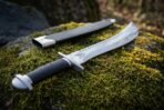 Short sword with upswept saw spine and steel pommel cap on a moss covered stone beside a black scabbard with steel throat