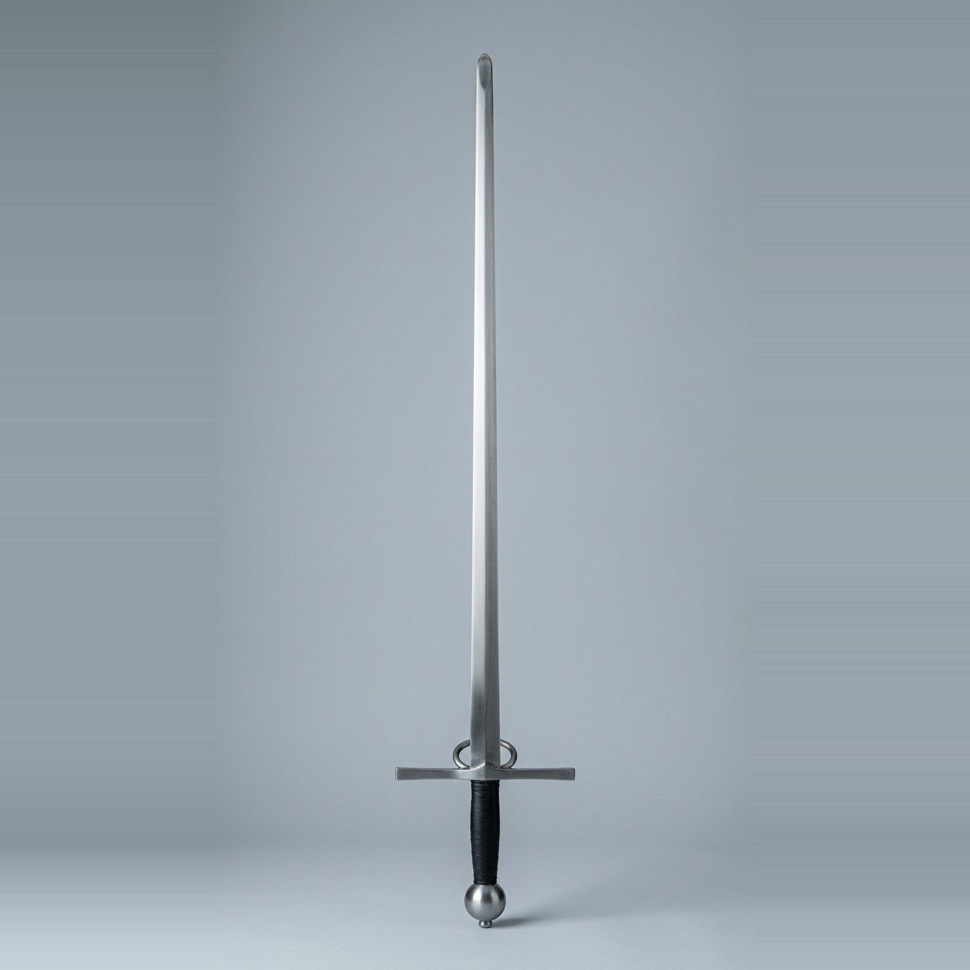 Ashgrimm side ring arming sword trainer Side ring arming sword trainer with two rings above a straight cross, a narrow unfullered blade, black leather grip and sphere pommel laid on a plain grey studio backdrop