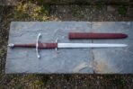 Side ring sword with burgundy leather grip and steel ring guard on a slate slab beside its burgundy scabbard, top view