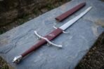 Side ring sword with raven stamped burgundy grip and faceted pommel lying diagonally on slate with its scabbard behind