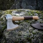 Side view of a chevron etched hand axe with square hammer poll and leather wrapped ash handle resting on a mossy stone