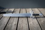 Side view of a Damascus sword with loop guard and disc pommel resting on weathered boards near its scabbard