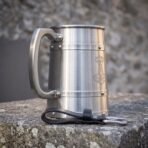 Side view of a stainless steel tankard with etched Thor hammer and riveted bands on a stone ledge with a black leather belt hanger