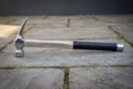 Side view of a steel claw hammer with rune line on a pale hickory handle, black rubber grip and steel end cap on paving