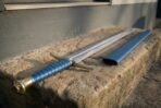 Side view of a steel sword with blue wrapped grip and brass pommel resting on a sunlit stone step beside its blue scabbard