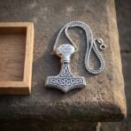 Silver finish Mjolnir pendant with a vegvisir crown and beaded edge laid flat on stone with its box chain and an oak box