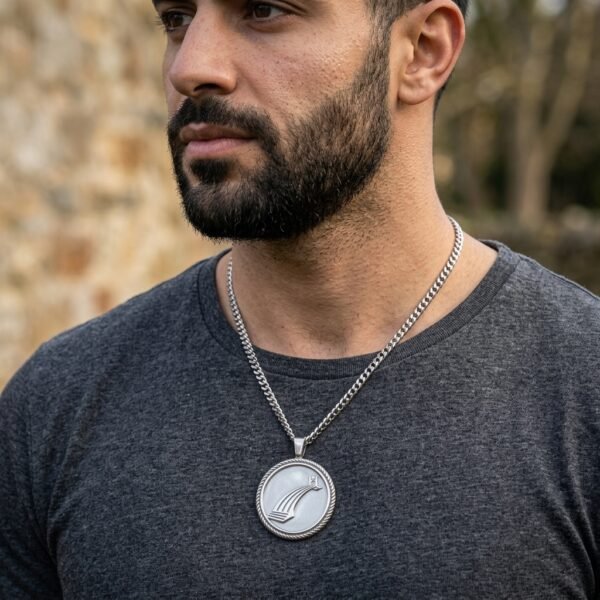 Silver tone bifrost pendant on a heavy curb chain worn over a grey shirt by a bearded man beside a stone wall