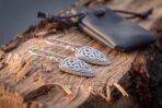 Silver tone Celtic shield earrings with hook wires lying on split wood in front of a grey leather pouch