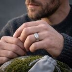 Silver tone rune ring with elder futhark band worn on a man's finger, hands clasped over a mossy rock with a suede pouch