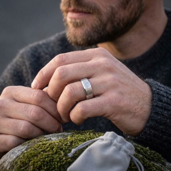 Silver tone rune ring with elder futhark band worn on a man's finger, hands clasped over a mossy rock with a suede pouch