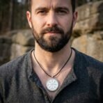 Silver tone vegvisir pendant on a black leather cord worn by a bearded man in a grey shirt in front of a stone wall