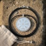 Silver tone vegvisir pendant on a black leather cord with lobster clasp laid flat on sandstone, drawstring linen pouch alongside
