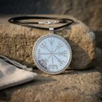 Silver tone vegvisir pendant with twisted rope bezel and black leather cord propped on a sandstone block beside a linen pouch