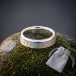 Silver tone Viking rune ring with an Elder Futhark band cut into a brushed centre panel and polished bevelled edges standing on wet moss beside a grey suede pouch