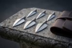 Six Damascus steel broadhead arrowheads laid in two rows on a weathered stone slab next to a rolled brown leather case