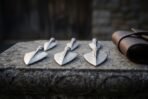 Six Damascus steel socketed broadhead arrowheads standing point up on a stone ledge beside a rolled brown leather case