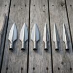 Six hand forged viking spearhead blades in graduated sizes with socketed collars and rune marks laid out on weathered grey decking beside a rolled canvas tool roll